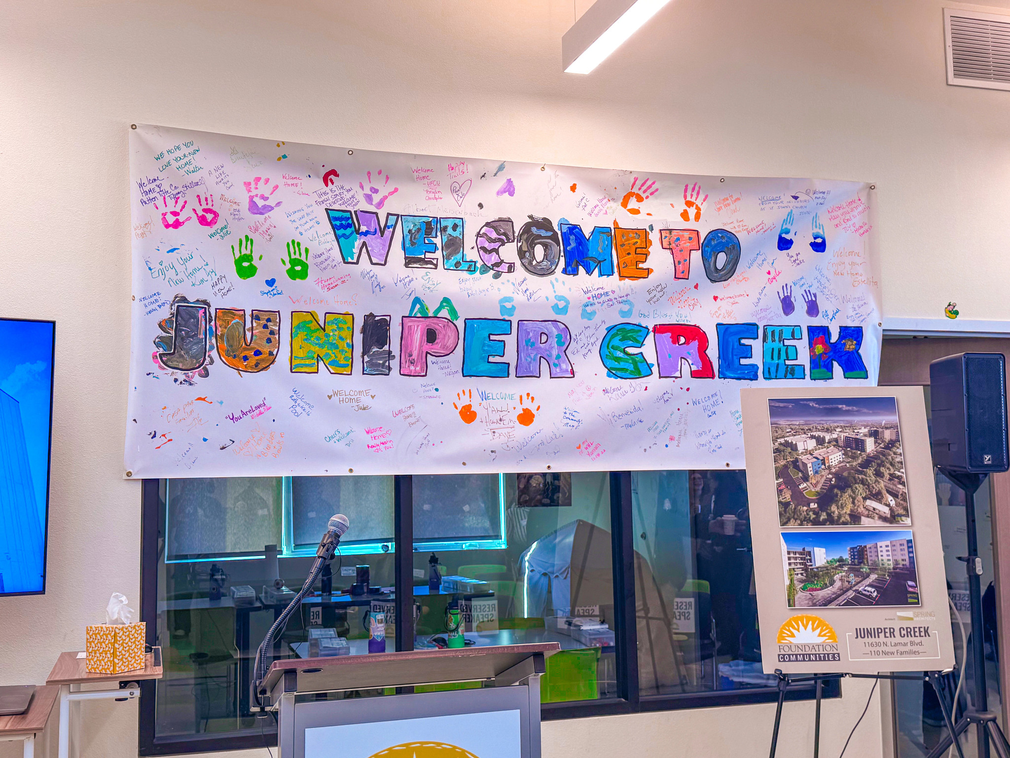 “Welcome to Juniper Creek” banner decorated with colorful handprints and handwritten messages from residents, displayed above a podium and microphone during the Juniper Creek grand opening event. An easel sign shows renderings of the 110-unit affordable h