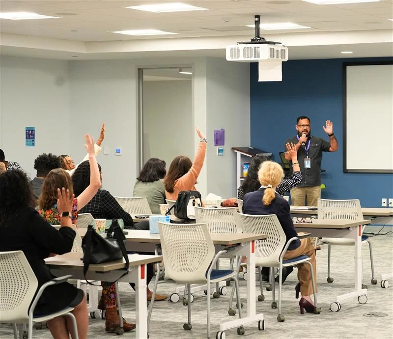 A speaker addresses an audience in a conference room. Several attendees raise their hands, engaging with the presentation. The atmosphere is interactive.