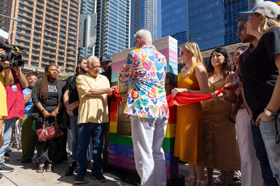 A crowd surrounds two men shaking hands in front of a rainbow-colored brick marker