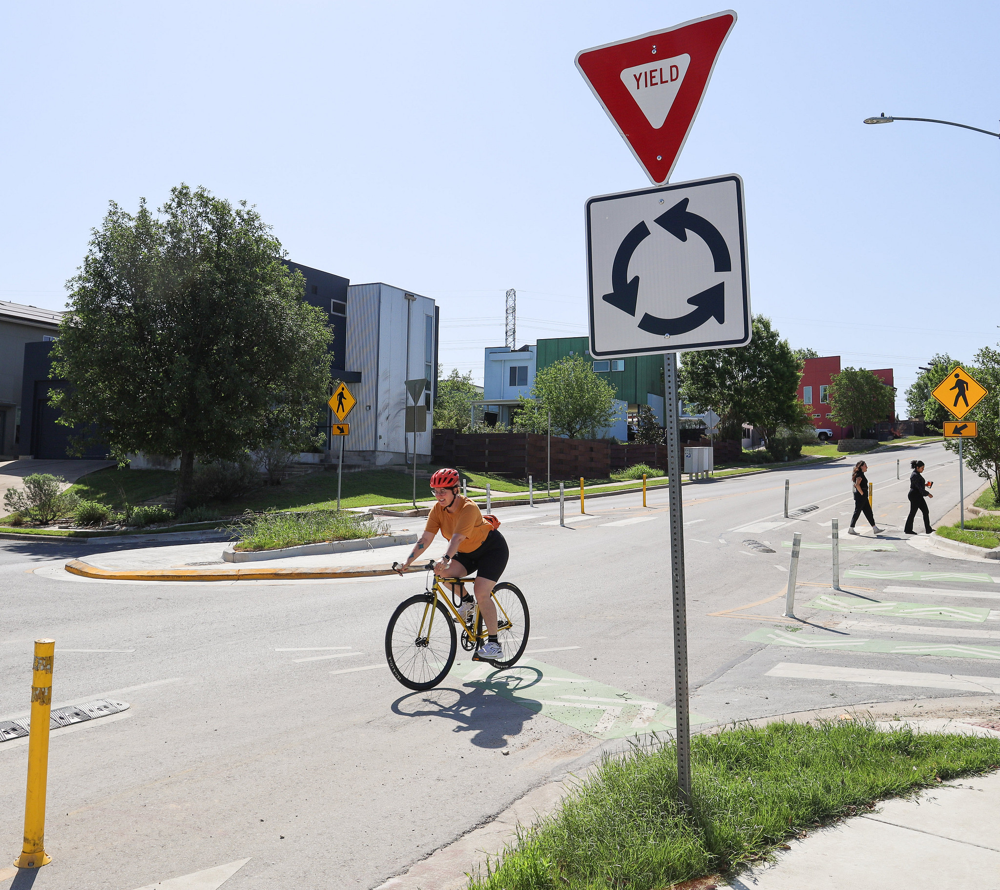 Person rides bicycle while two others cross the street at an intersection with a roundabout.