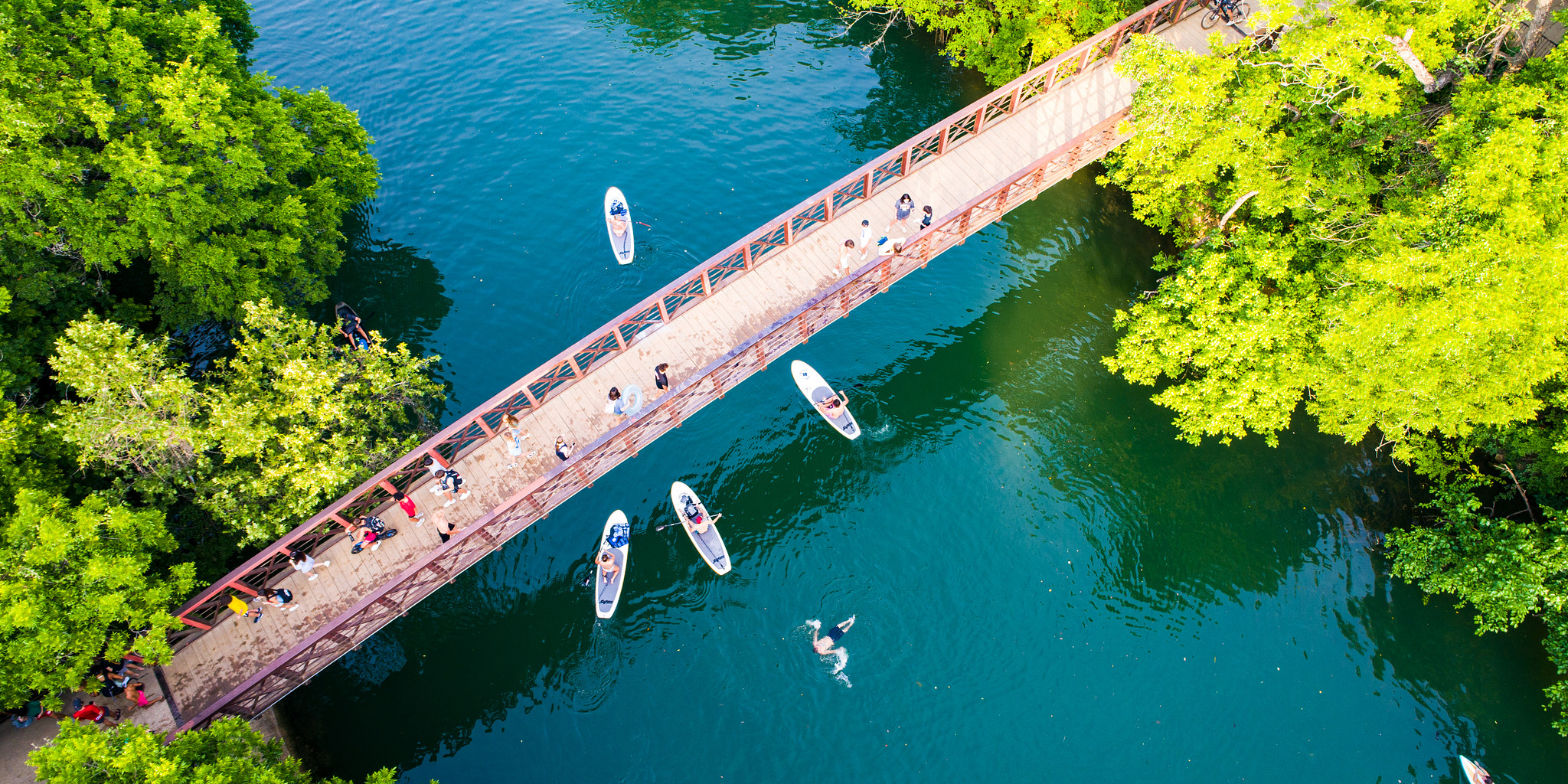 A bridge with people on it over a beautiful blue waterway with people swimming and paddle boarding.