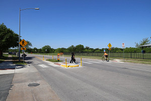 People cross a street with a pedestrian crossing island. A bicyclist rides in a two-way cycle track.