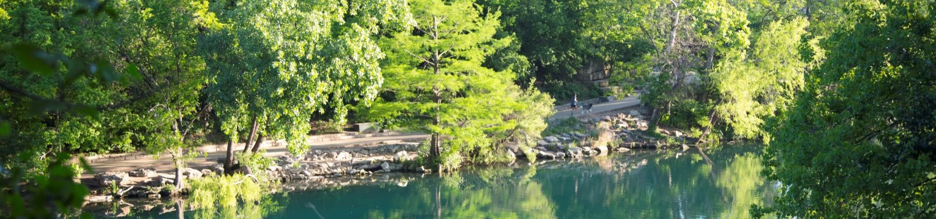 Person walking a dog on a paved trail by a river lined with trees