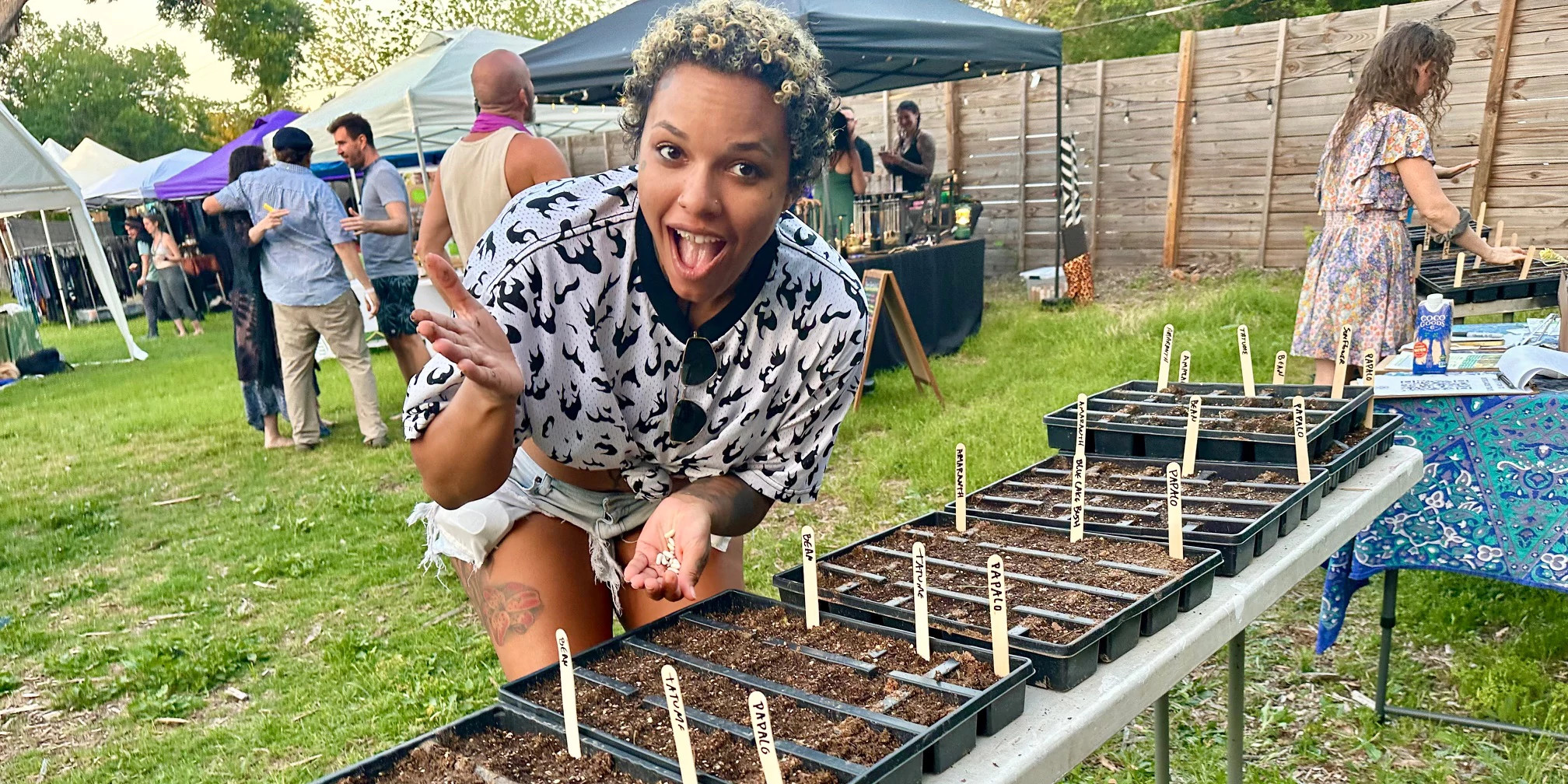 People gather at an event with several tents, with a person in the foreground smiling and gesturing to some trays of planted seedlings.