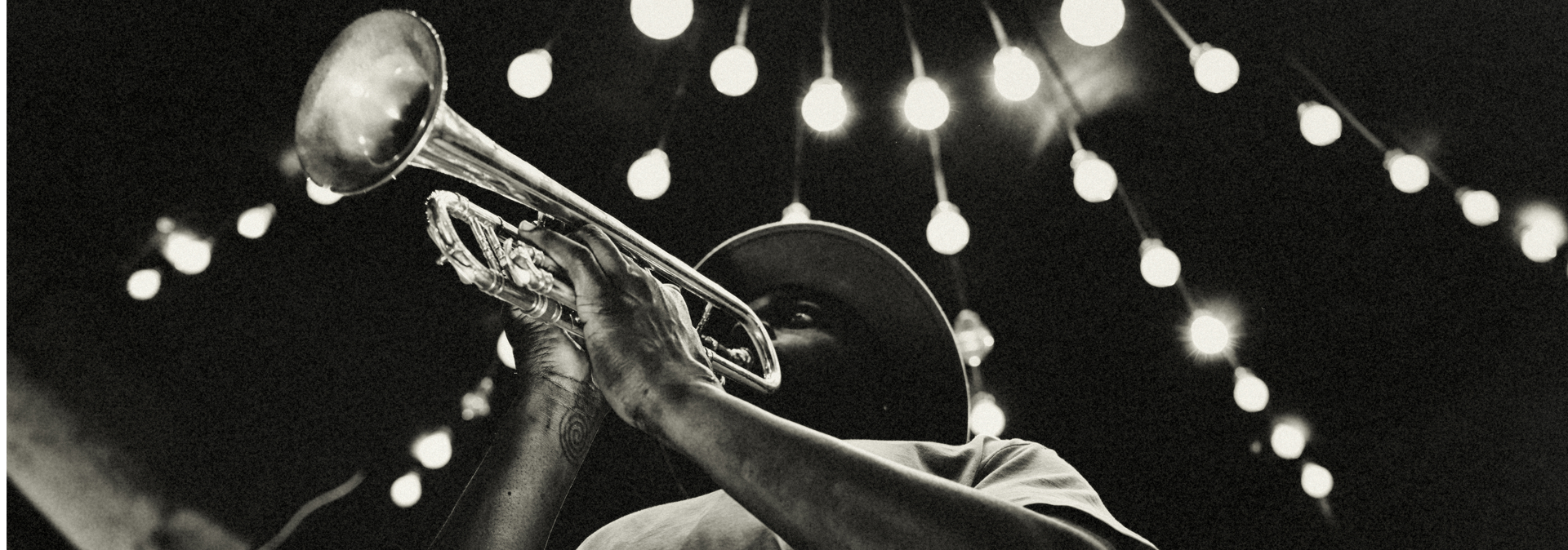 A black and white photo of a musician playing the trumpet under string lights.