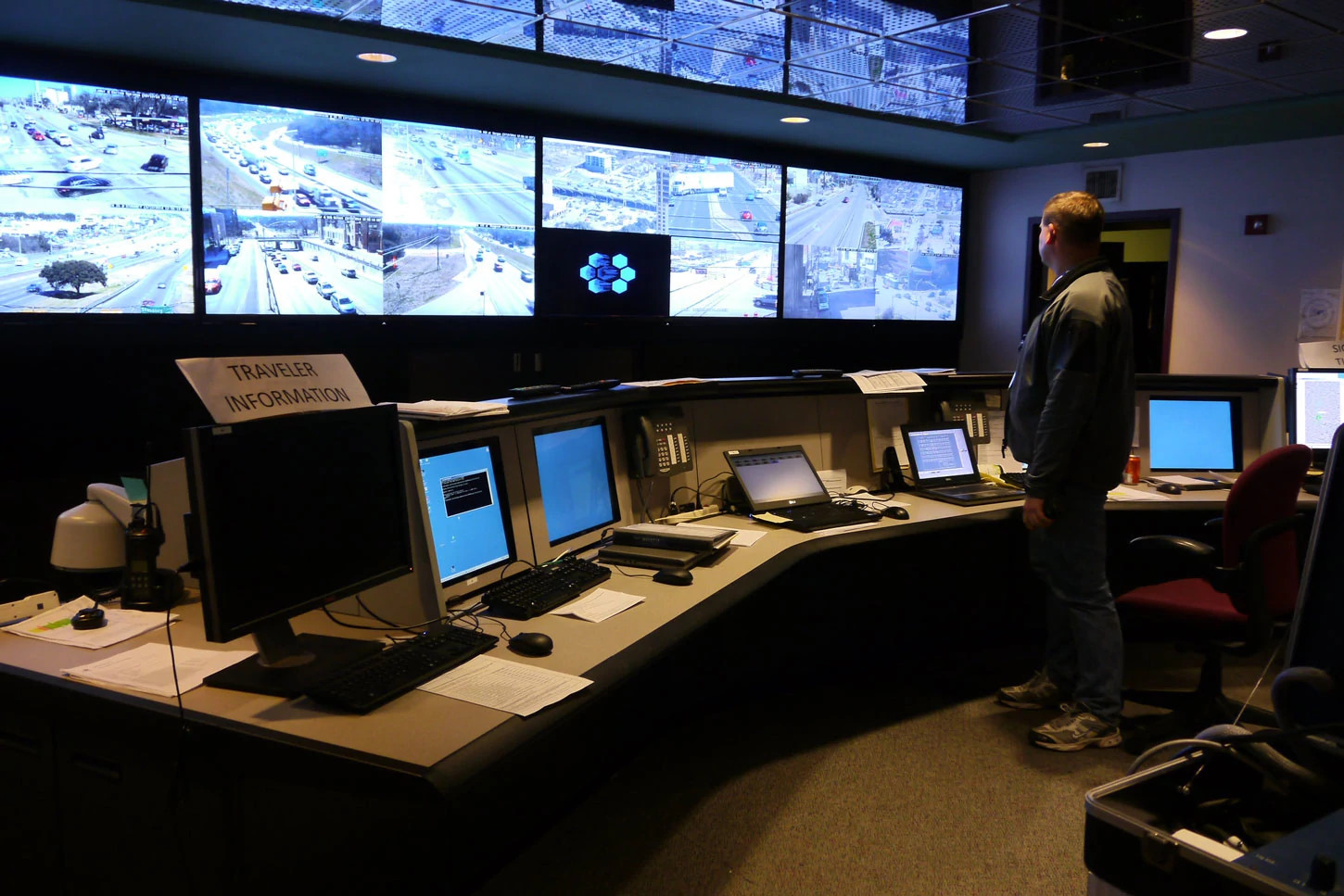 A man standing in a dark room looking at a wall of monitors showing cars around town