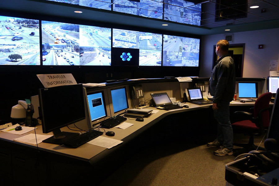 A man standing in a dark room looking at a wall of monitors showing cars around town