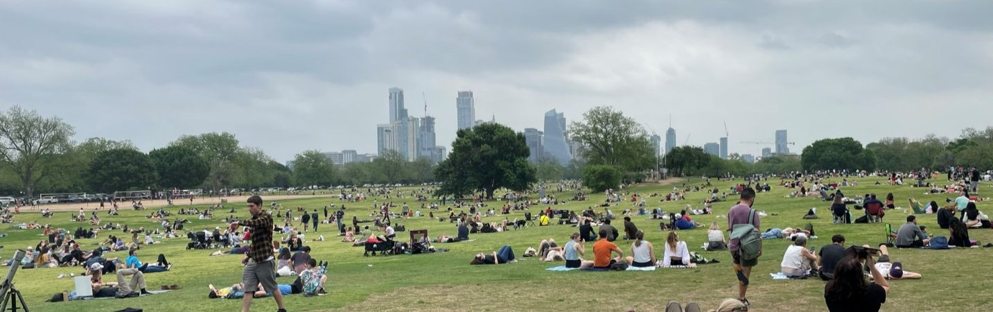 People relaxing on a grassy lawn in Zilker Park with a city skyline in the distance.