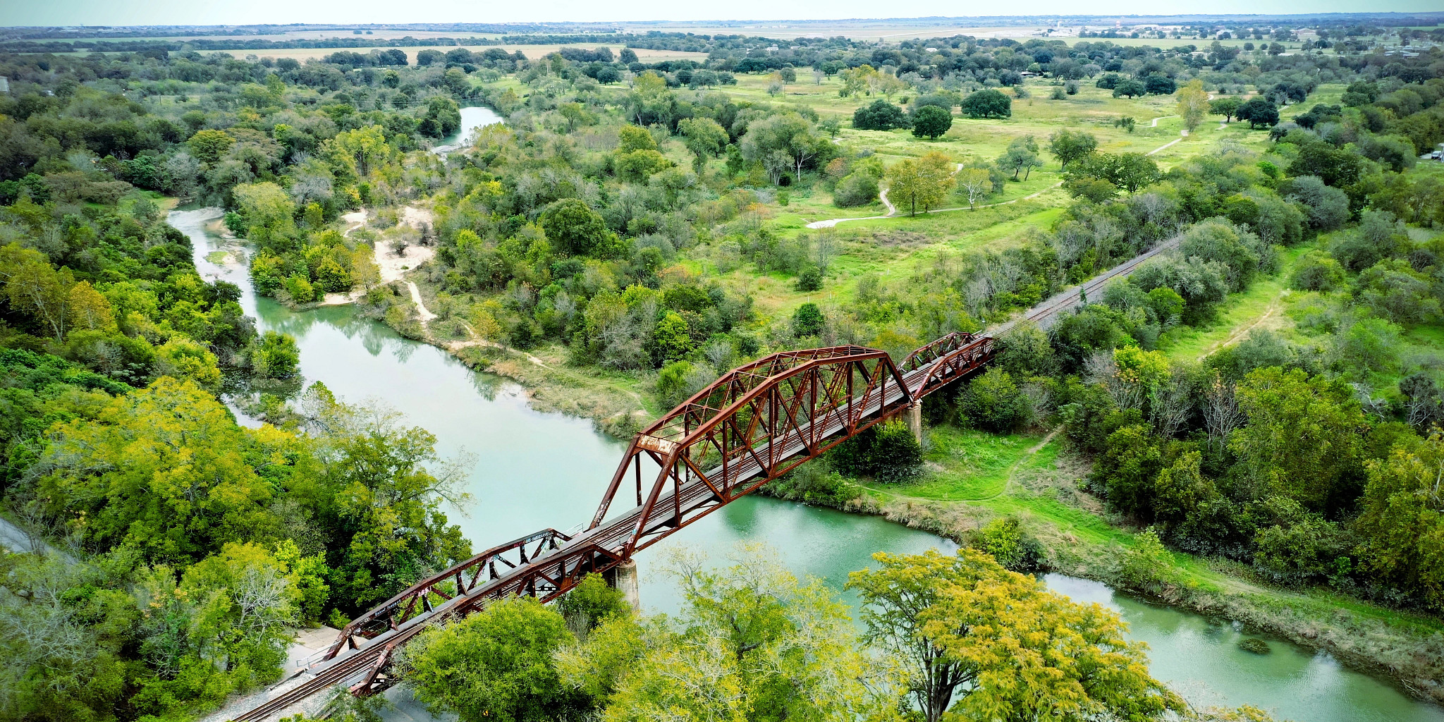 Photo of a bridge over a clear river with green trees all around and not many buildings