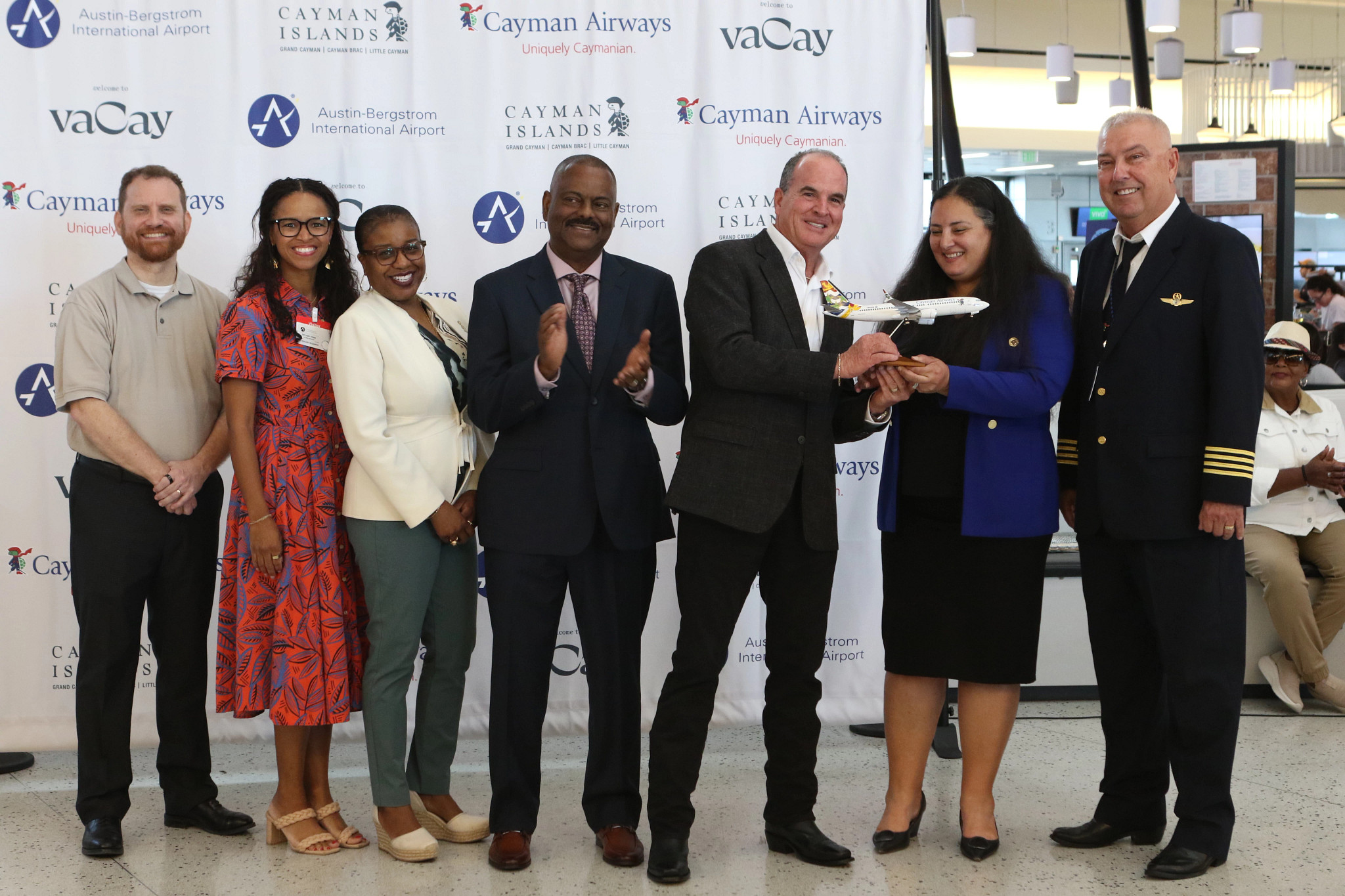 Group photo at Austin-Bergstrom International Airport celebrating Cayman Airways service. Seven individuals stand in front of a branded backdrop featuring AUS, Cayman Airways, and Cayman Islands logos. Two people in the center hold a model Cayman Airways 