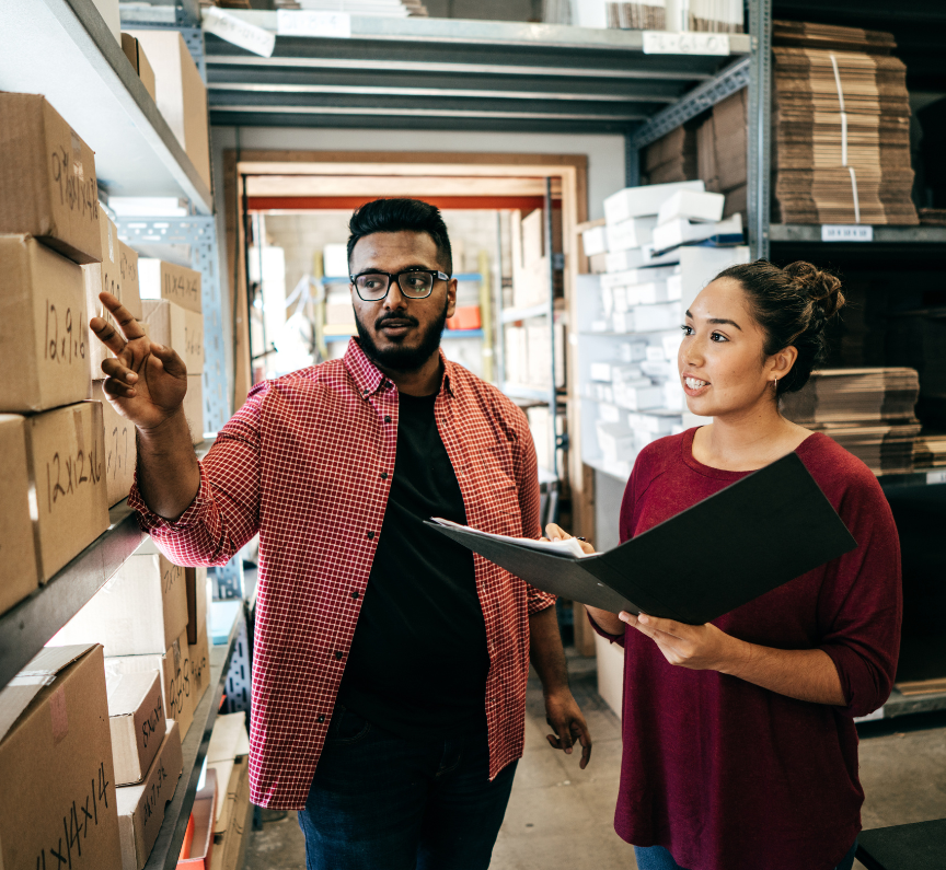 two individuals in a distribution warehouse, pointing and discussing a stack of cardboard boxes