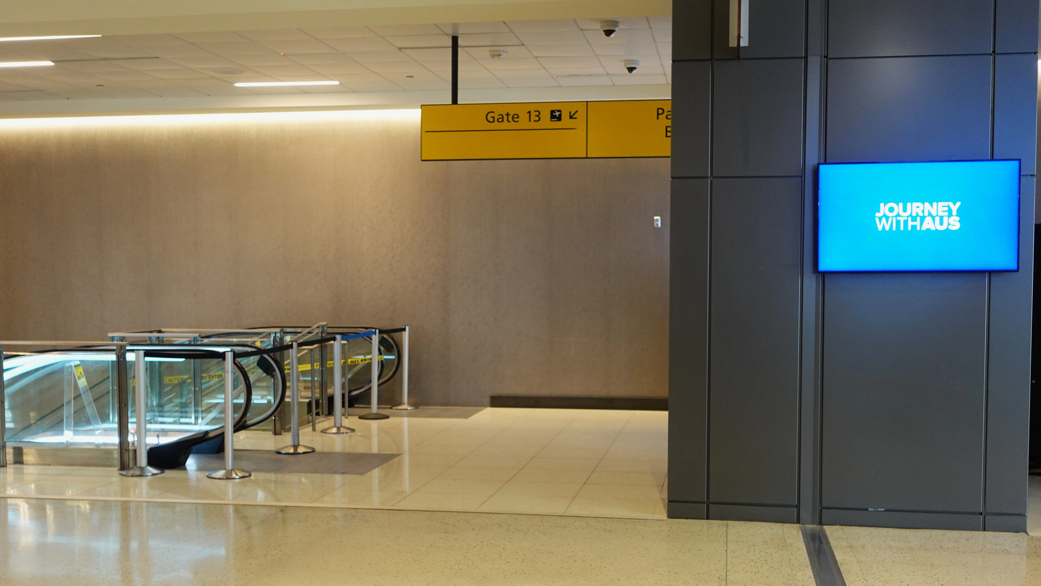 A picture of the entrance to the AUS gate 13. There is a television screen on the right, and escalators to the left, which lead down to the hold room of the gate 13.