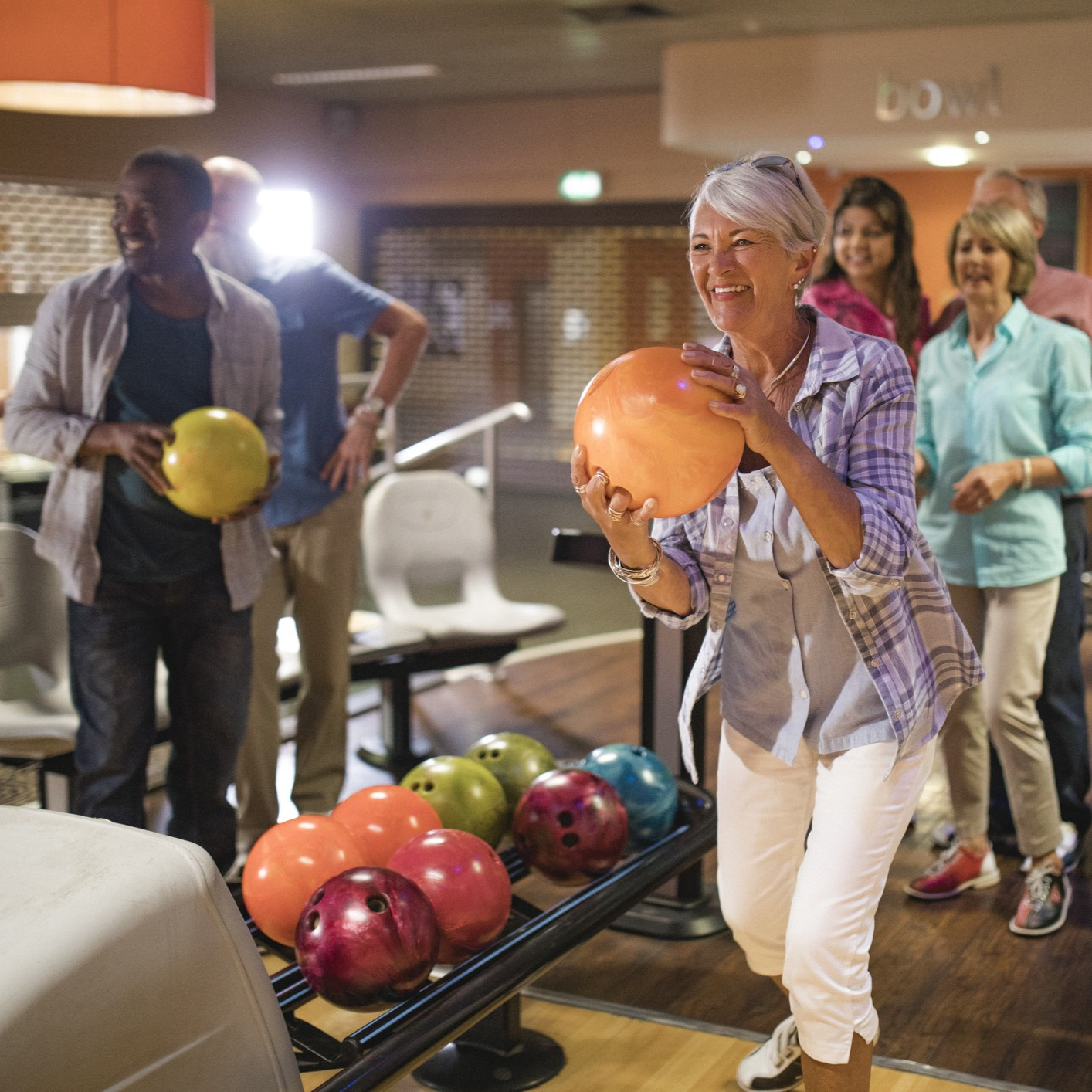 Group of seniors at a bowling alley, five senior friends can be seen in the background whilst a woman takes her turn.