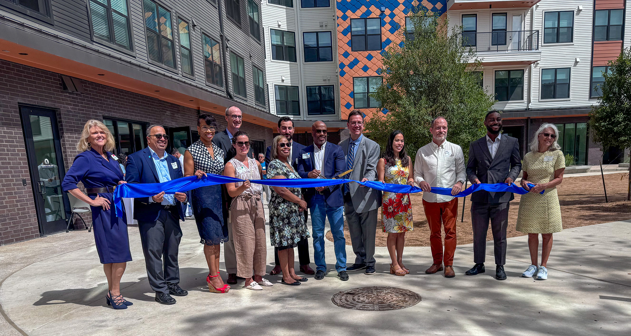 Group of people standing outdoors in front of Seabrook Square I, holding a long blue ribbon for a ribbon‑cutting event.