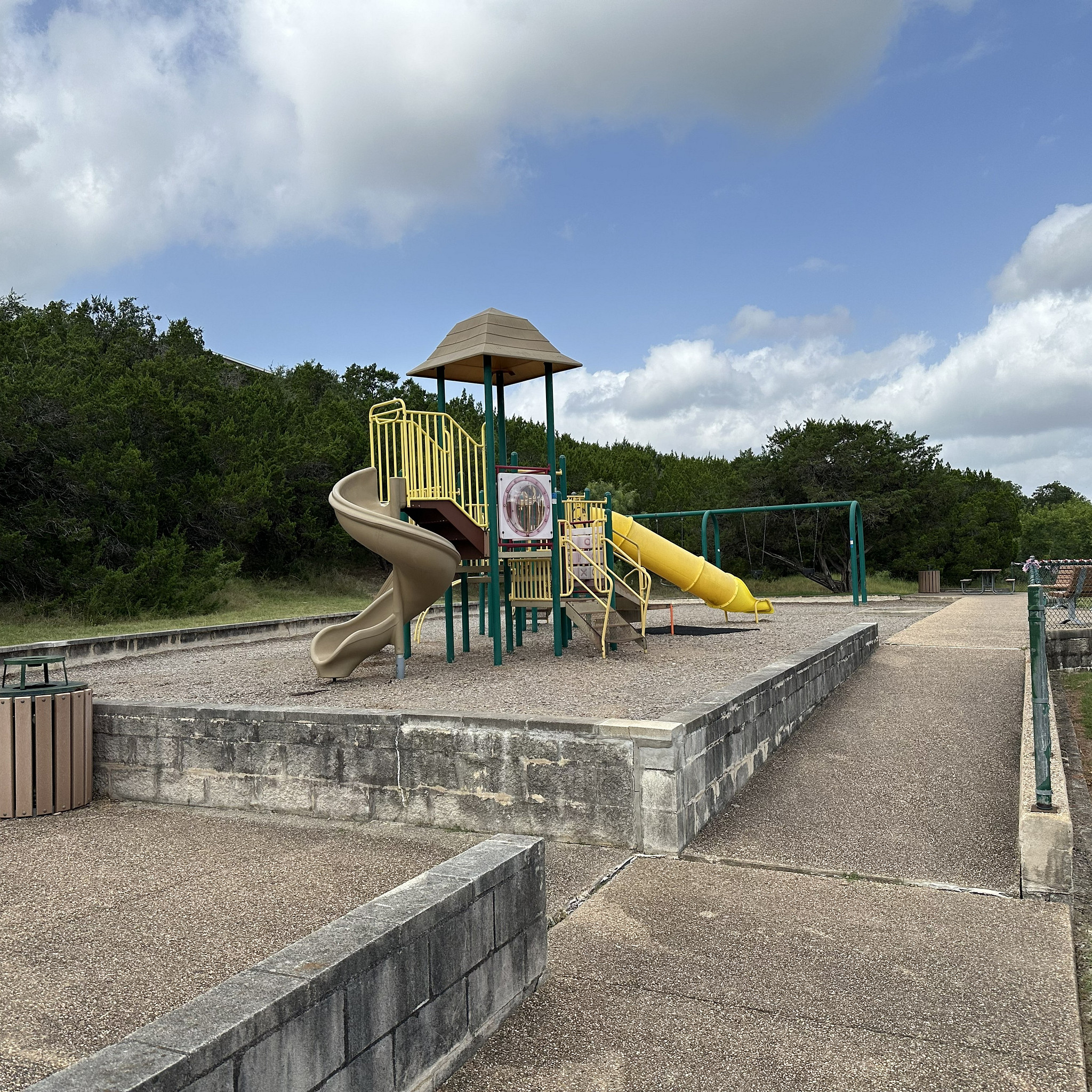 Play structure at Piney Bend surrounded by concrete sidewalks and retaining wall, trees in the background