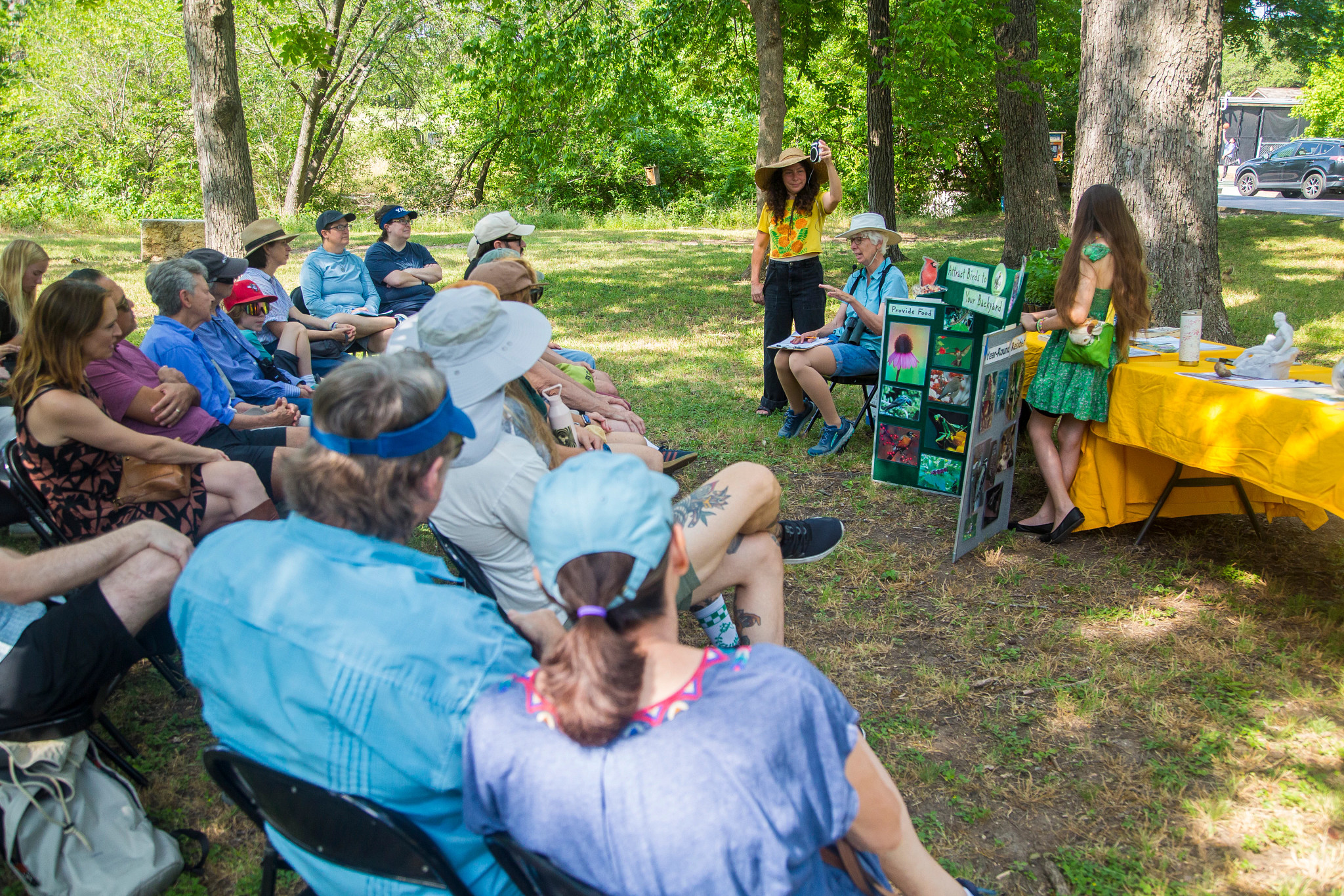 Elderly woman sitting in front of a tri-fold speaking to an audience in a backyard