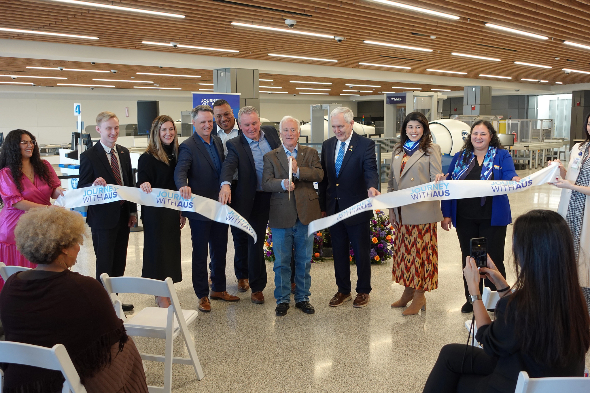 A group of airport and city leaders stand inside a bright, modern terminal area holding a ribbon that reads “Journey With AUS” during a ribbon-cutting ceremony. They smile for photos in front of new security screening equipment, marking the opening of a newly expanded checkpoint space.