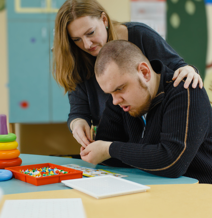 Woman assists young adult student with a sorting task at a schoolwork table.