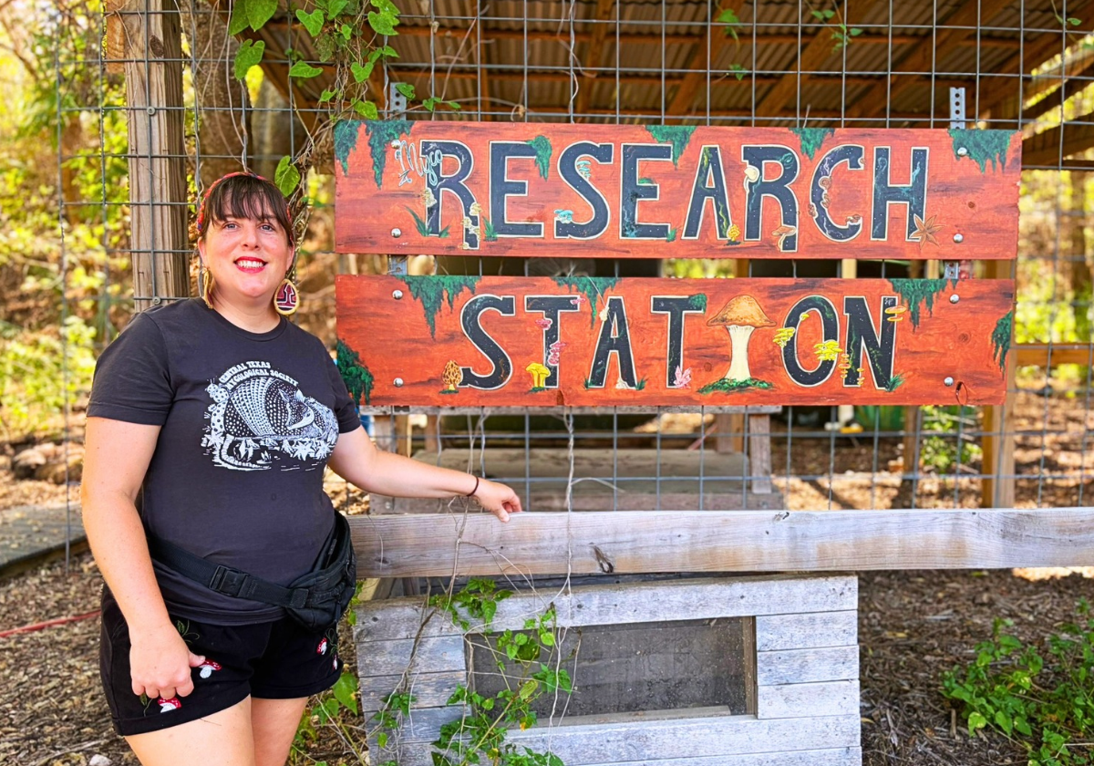 Angel stands in front of a hand-painted sign reading Research Station. Illustrations of mushrooms pop out from the letters.