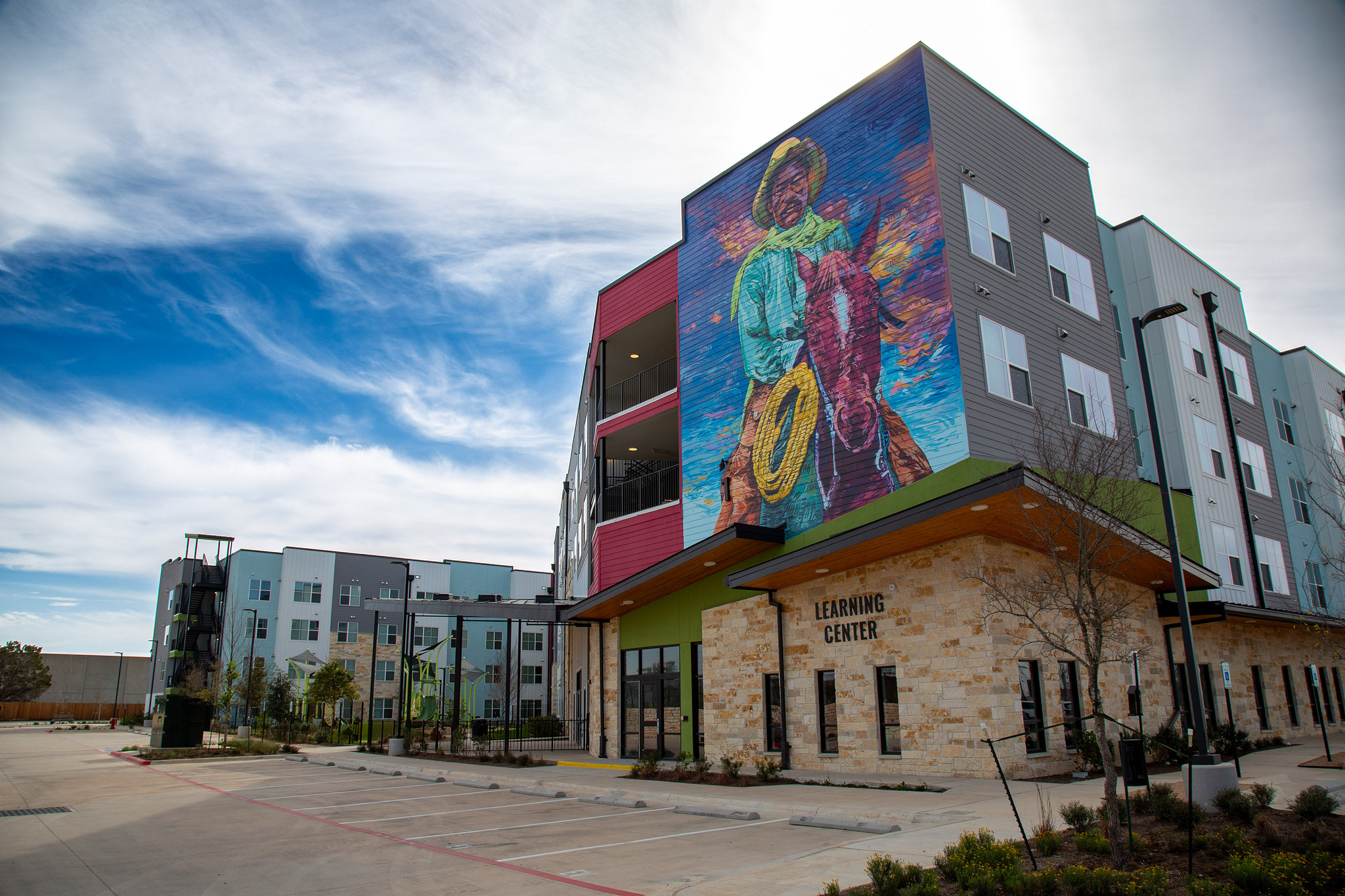 Image shows a close-up view of the leasing office for Norman Commons with stone on the first floor and a large mural of an African American East Austin Cowboy. The mural scales multiple floors above the office. Other multi-story buildings can be seen in t