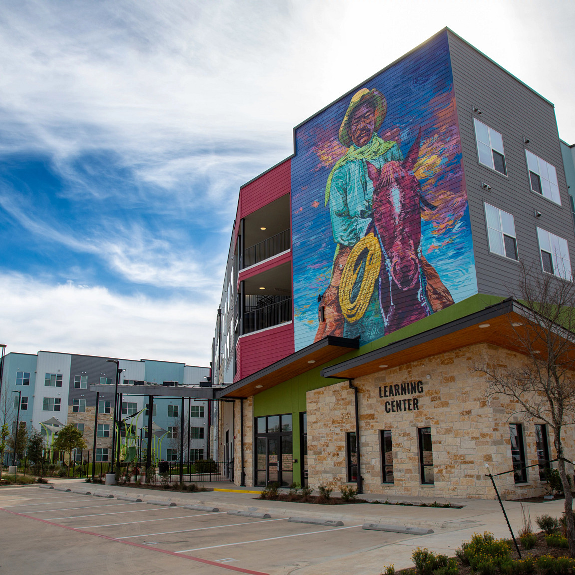 Image shows a close-up view of the leasing office for Norman Commons with stone on the first floor and a large mural of an African American East Austin Cowboy. The mural scales multiple floors above the office. Other multi-story buildings can be seen in t