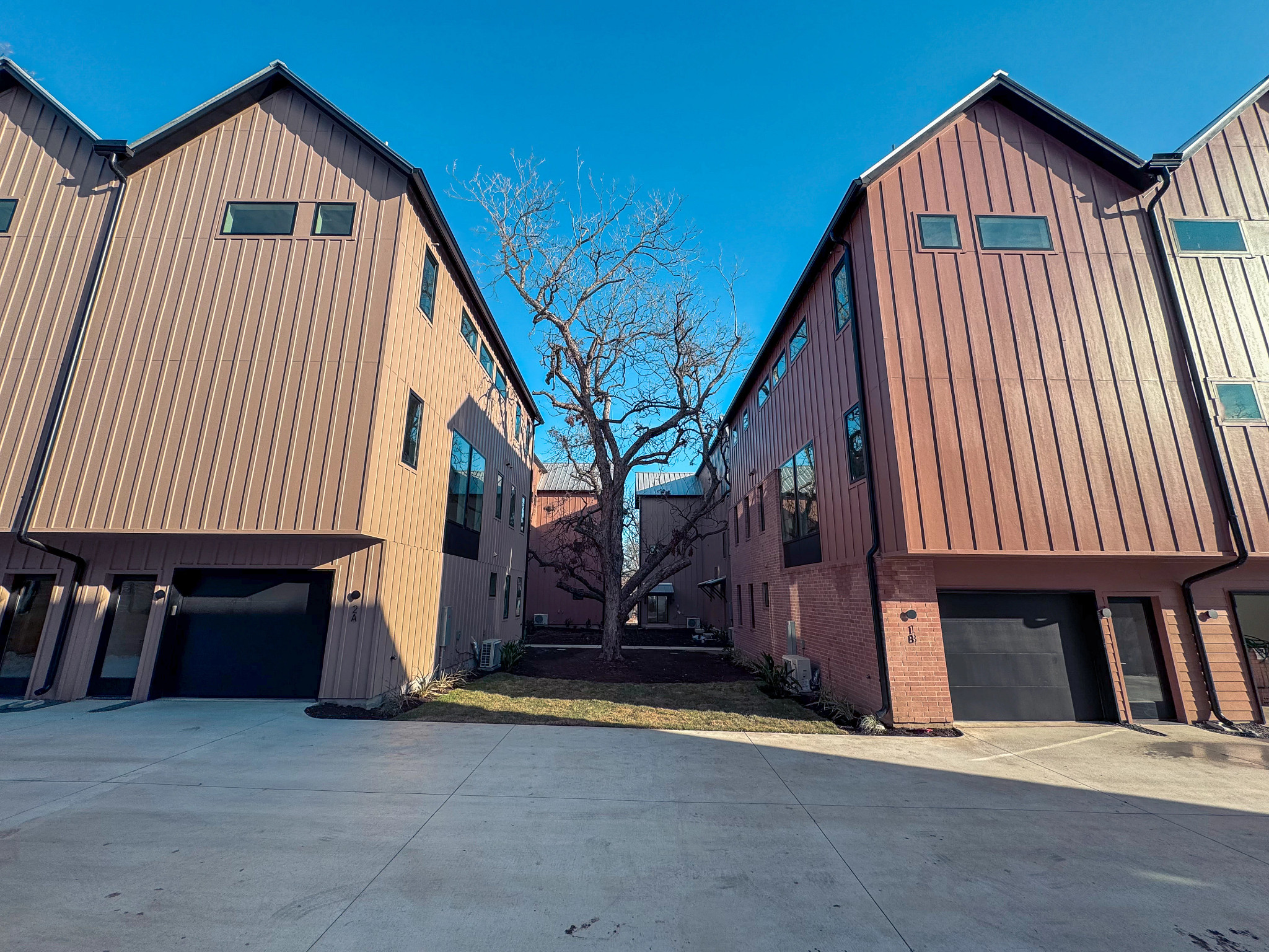 Two modern townhouse buildings face each other across a concrete driveway, with a leafless tree centered between them under a clear blue sky.