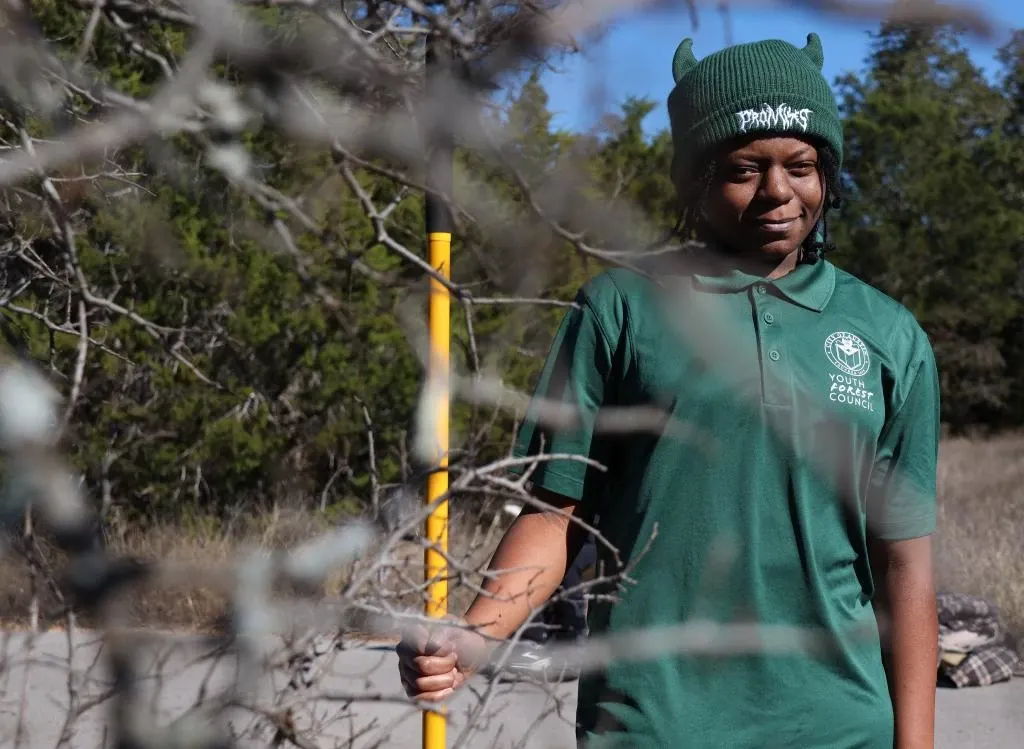 Jalinda stands in a Youth Forest Council shirt holding a yellow tool near a bunch of trees.