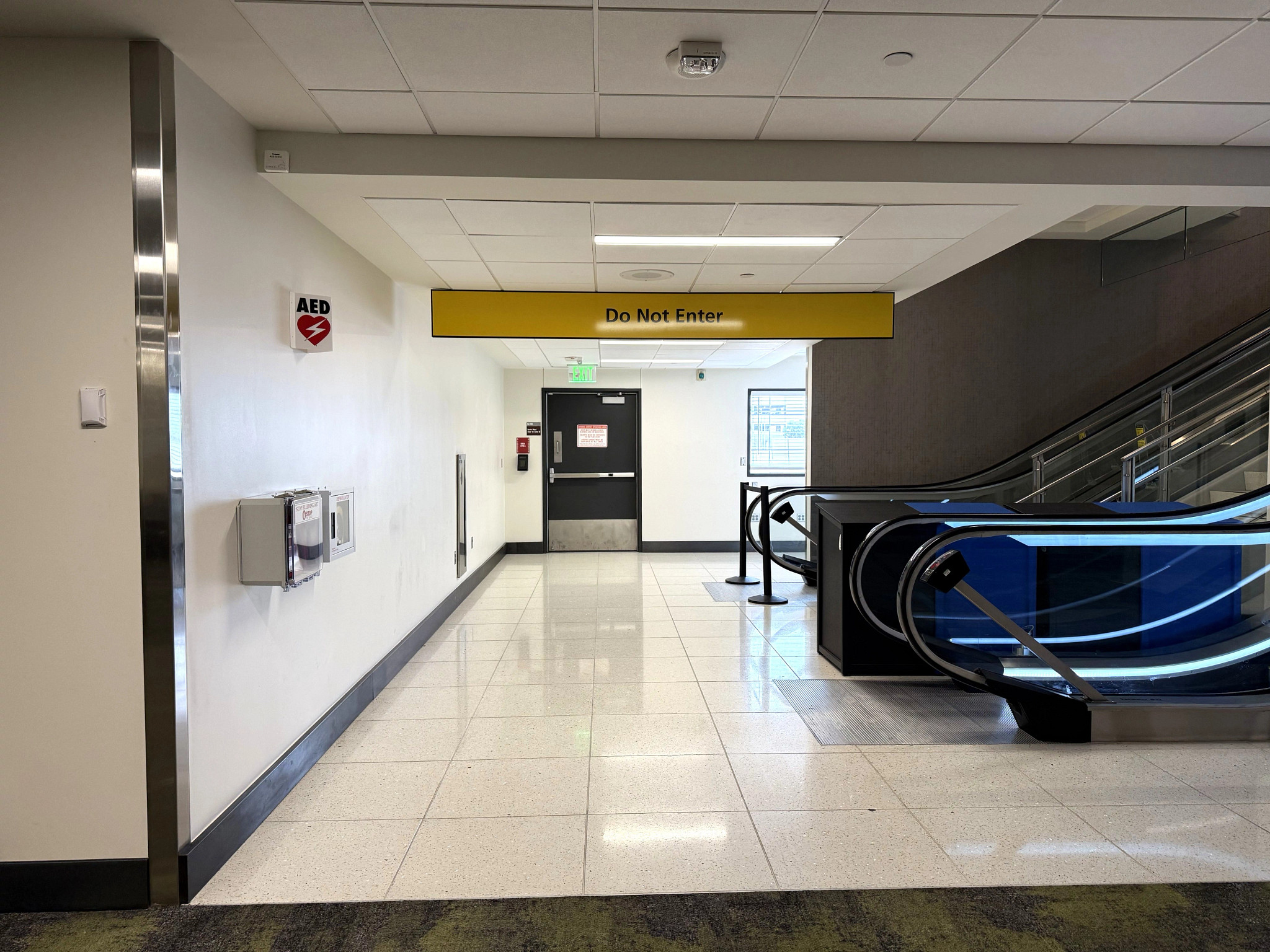 A picture of the bottom to the AUS gate 13 escalator leading to the hold room area. There are escalators on the right. There is a sign on the ceiling that reads Do Not Enter. There is a door in the middle of the space and an AED machine on the right.