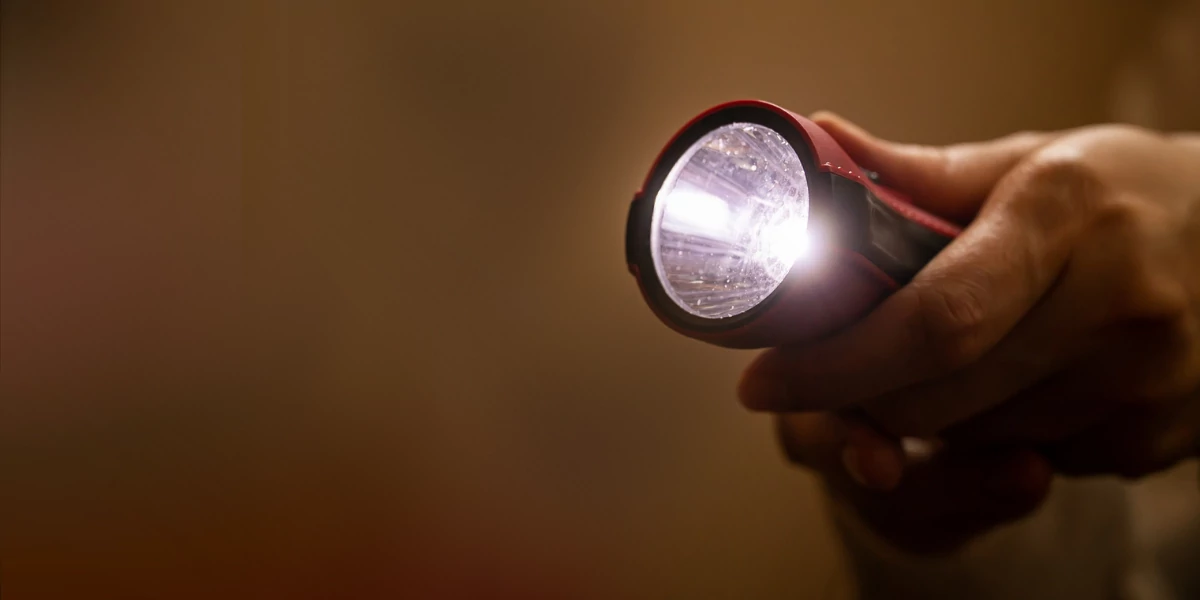 Closeup of person's hand holding a lit flashlight with a blurry dark brown background.