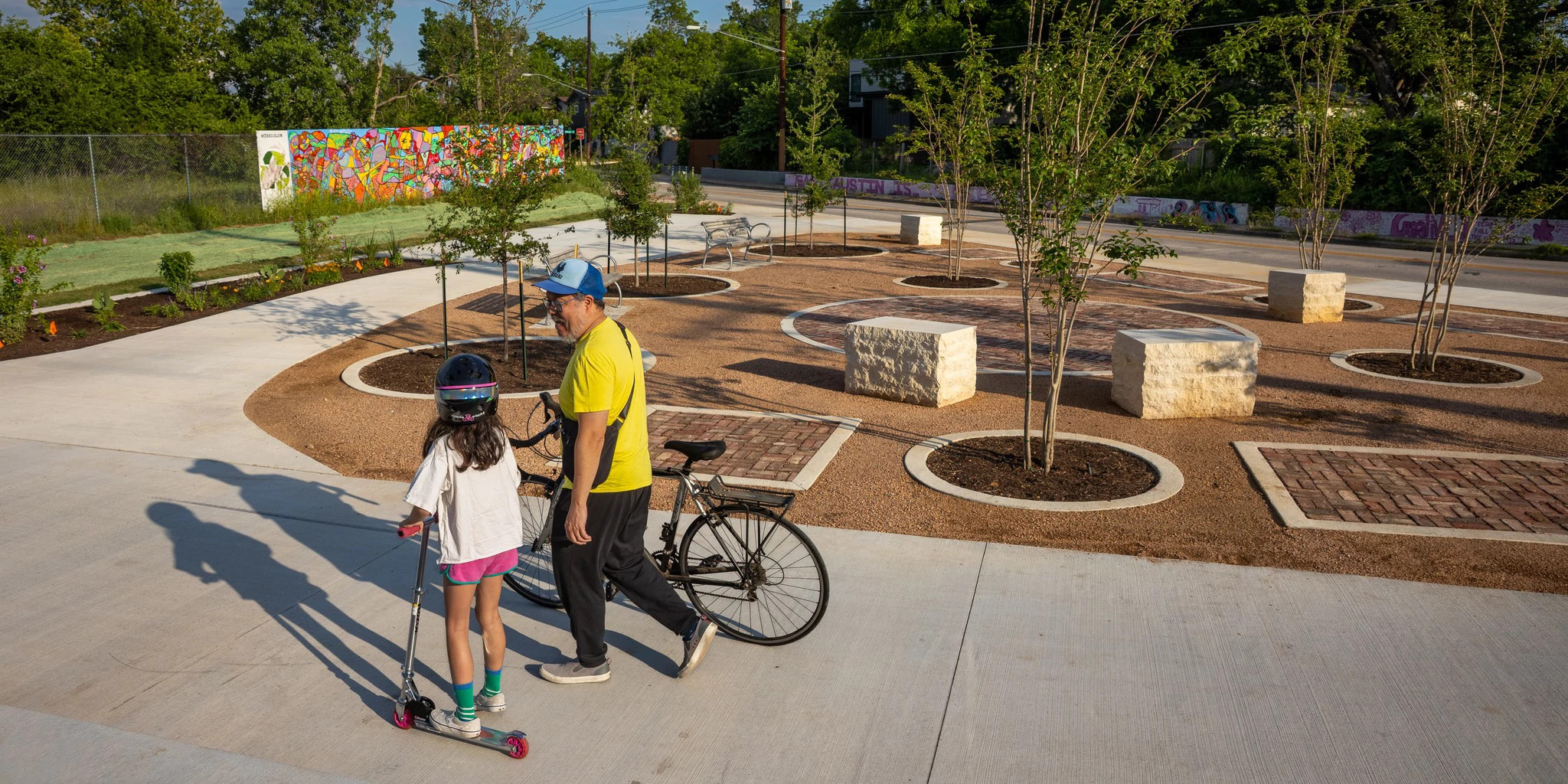 A person with a younger child using a scooter and bicycle moving through a park-like space with newly planted trees.