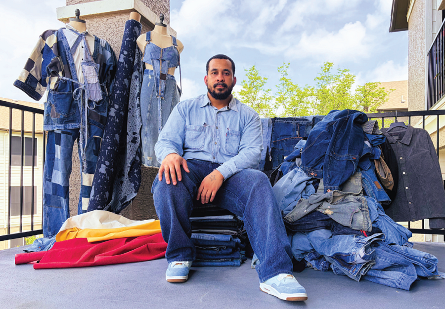 Billy sits on a pile of folded denim, surrounded by piles of denim, fabric bolts, and several of his recycled denim outfits.
