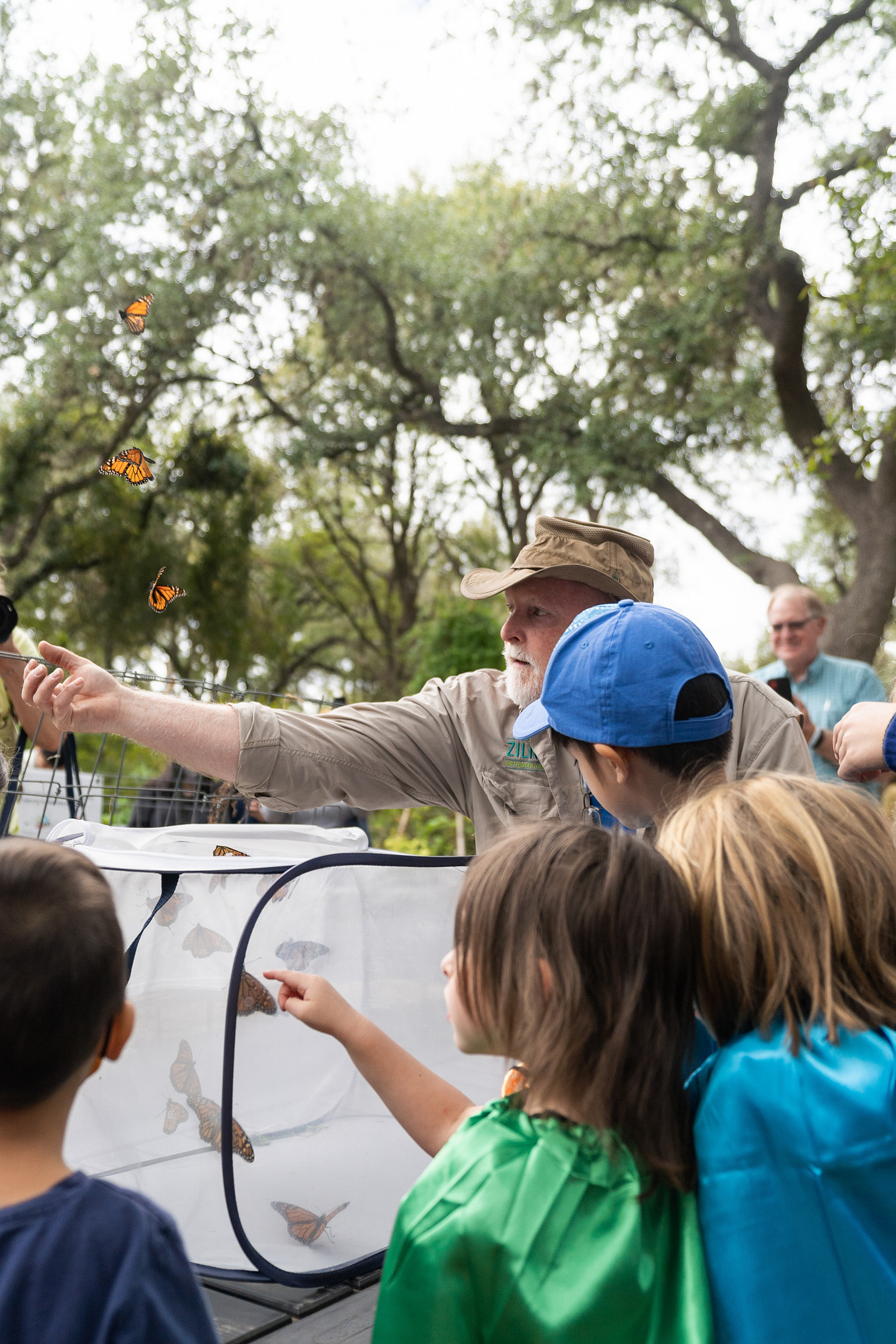 Man with arm outstretched helping butterflies out of net enclosure surrounded by kids watching. 