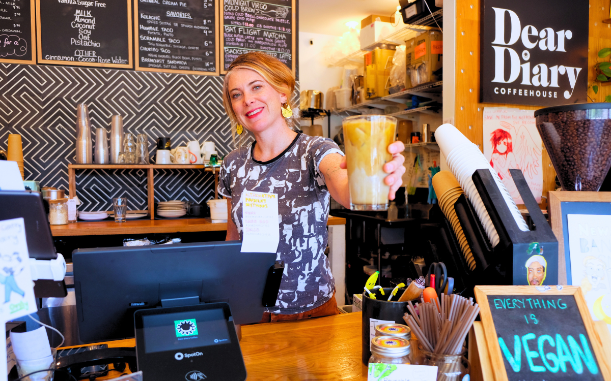 Litsa stands behind the bar at Dear Diary Coffee. She is smiling and holding out an ice latte.