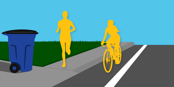 A person jogging on a sidewalk. A recycling cart is placed at the edge of a driveway, close to the sidewalk and road but not obstructing it. A cyclist rides a bicycle in the bike lane. 