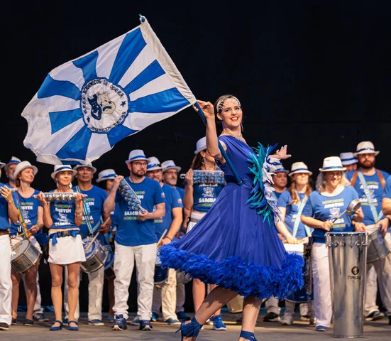An image of a Samba group on stage all wearing blue. Emphasis is on a person holding a flag.