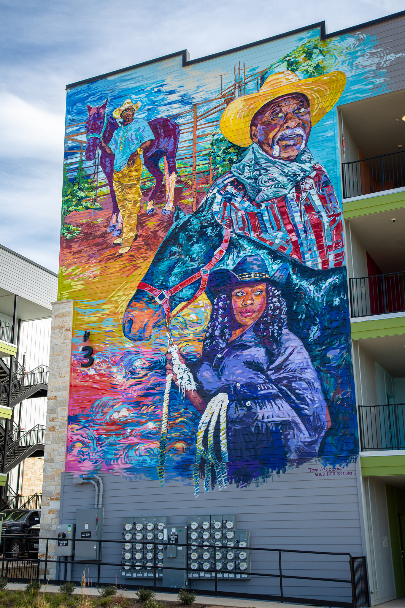 Image shows a close-up view of one of the Norman Commons buildings, which has a huge mural by Austin artist Toni Ardizzone depicting black cowboys, members of the East Austin Trail Riders in beautiful colors of blue, yellow, purple and red. The mural is p