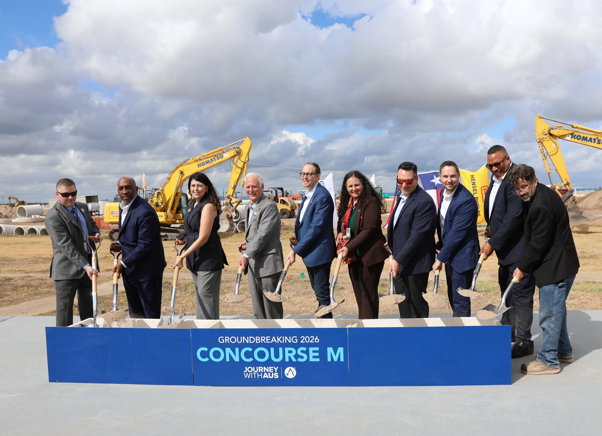 A group of ten people stand in a line at a construction site holding shovels for a groundbreaking ceremony. They are positioned behind a blue sign that reads Groundbreaking 2026 Concourse M Journey With AUS. Several large construction machines.