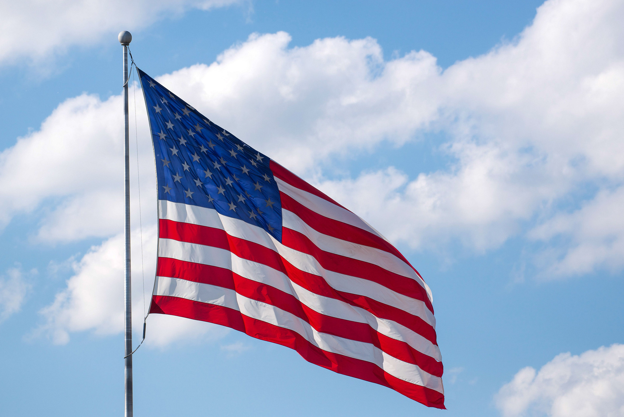 Large American flag on a flagpole waving in the winder under sunny blue skies and fluffy white clouds