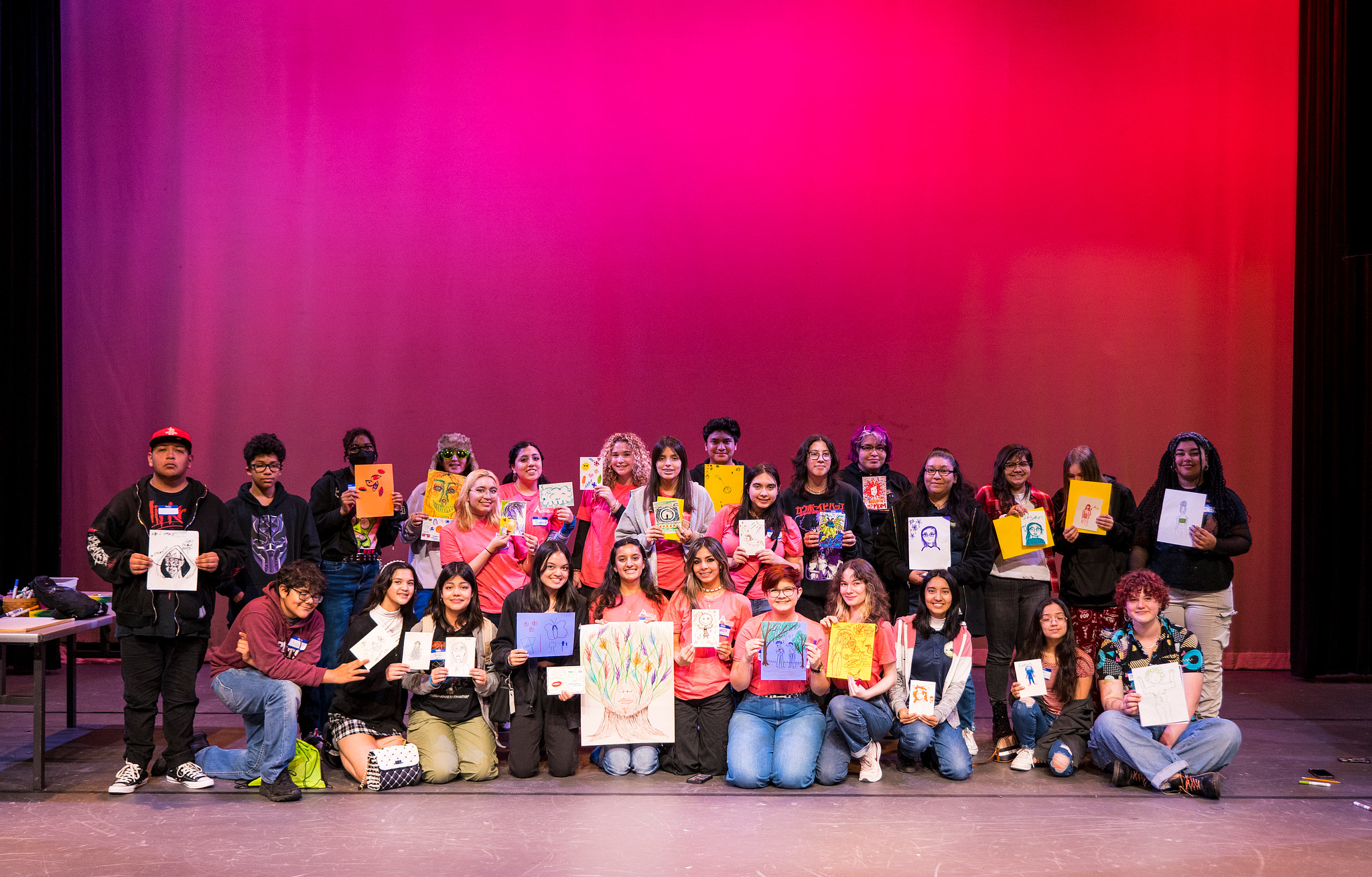 a group of 30 teens stand on a stage and holding up their artworks for a group photo