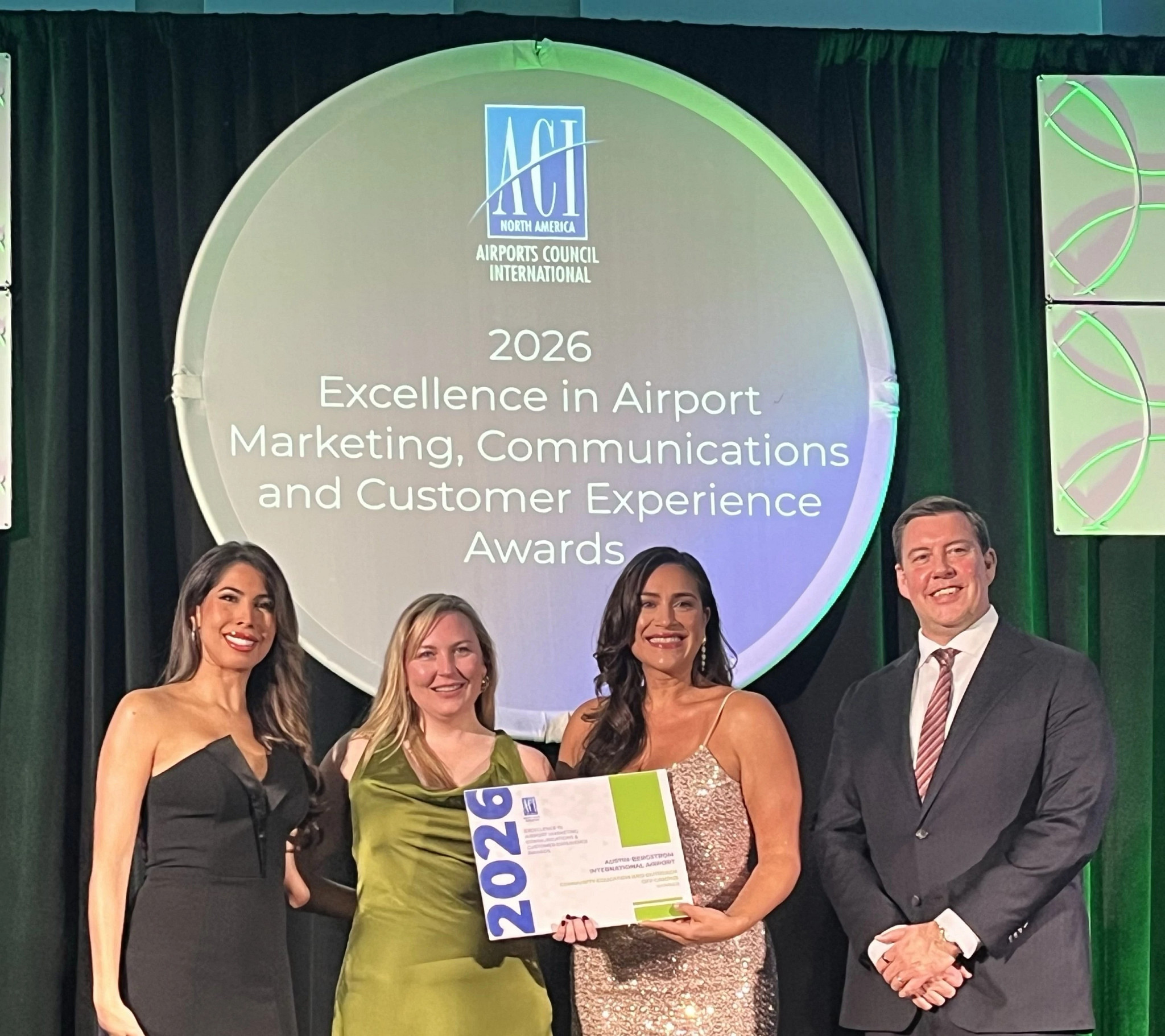 Four people standing on stage at an awards event, posing in front of a large sign that reads 2026 Excellence in Airport Marketing Communications and Customer Experience Awards. One person is holding a certificate, and all are dressed in formal attire.