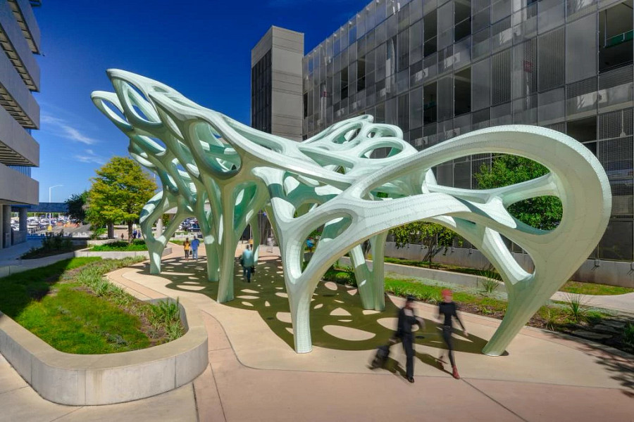 Outdoor public art installation featuring a large, pale green sculptural canopy with flowing, organic forms and cutout openings. The structure is supported by multiple curved legs and casts patterned shadows on the ground below. People walk beneath the sculpture along a landscaped pathway, with modern buildings and trees visible in the background under a clear blue sky.