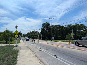 A person on a bicycle crosses a street with a crossing island and Rectangular Rapid-Flashing Beacon. The street also has protected bike lanes.