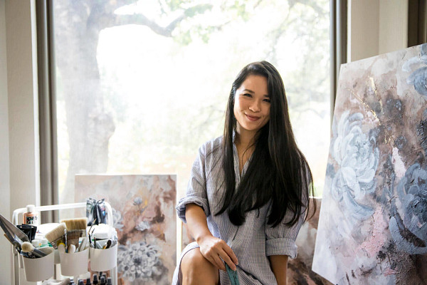 Picture of Lauren Hua sitting in front of paintings from Koi Pond, smiling at the camera