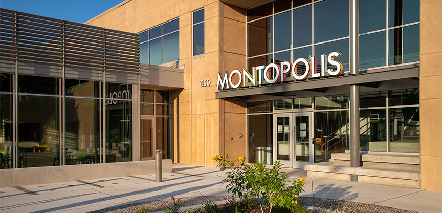 Exterior of the Montopolis Recreation and Community Center, a two-story tan building with large windows, native landscaping, and a wide concrete entrance.