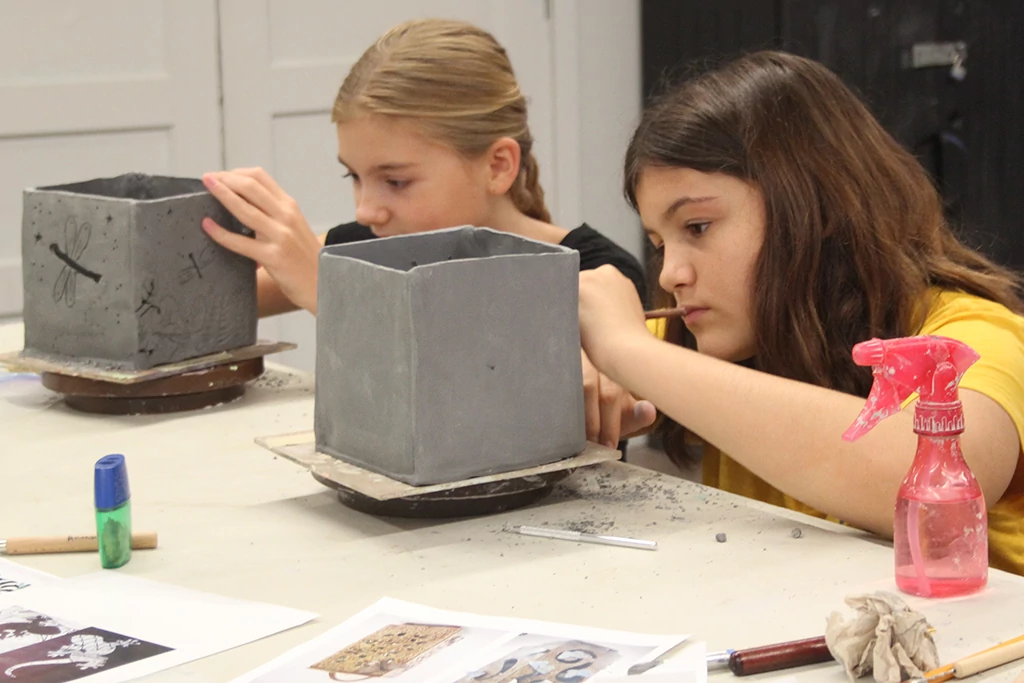 Two teen students concentrate on using tools to etch lines and shapes into the open-top cube-shaped ceramic projects on the worktable in front of them.