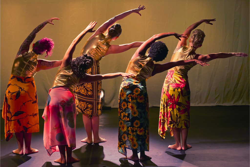 Five female dancer wearing gold tops and colorful skirts. They are standing in from of a curtain with their arms stretched towards the right of the screen.