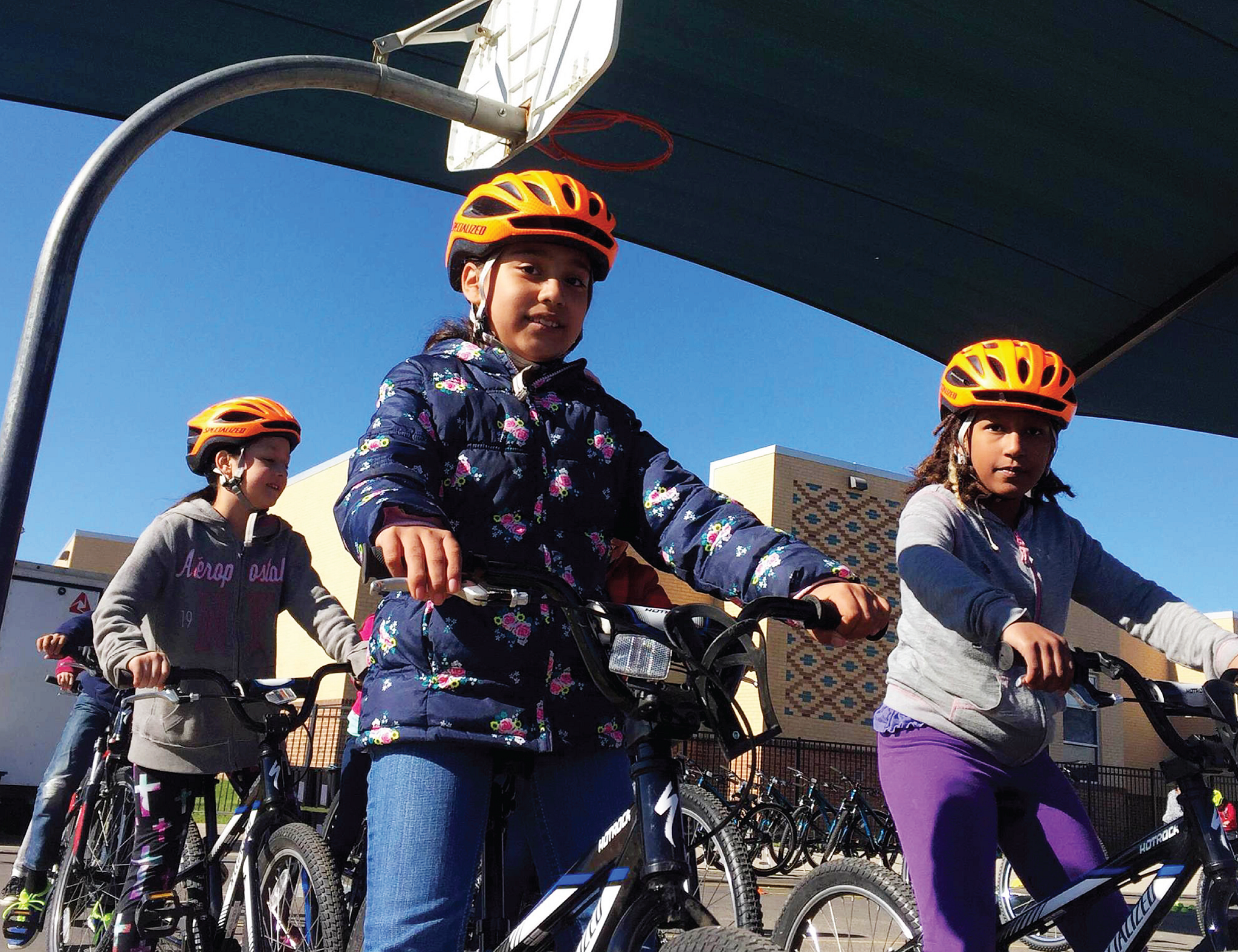 A group of students pause on bicycles to smile at the camera. They are wearing bright colored helmets and riding on a blacktop at Lee Elementary School.