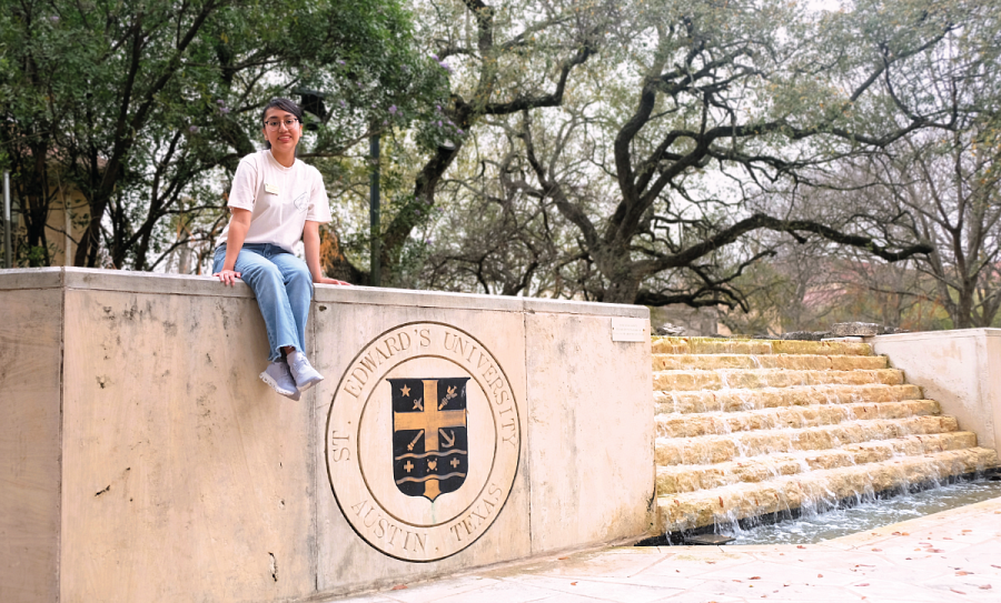Emily Hernandez-Corona smiles sitting on a stone wall with the St. Edward's emblem engraved in it.