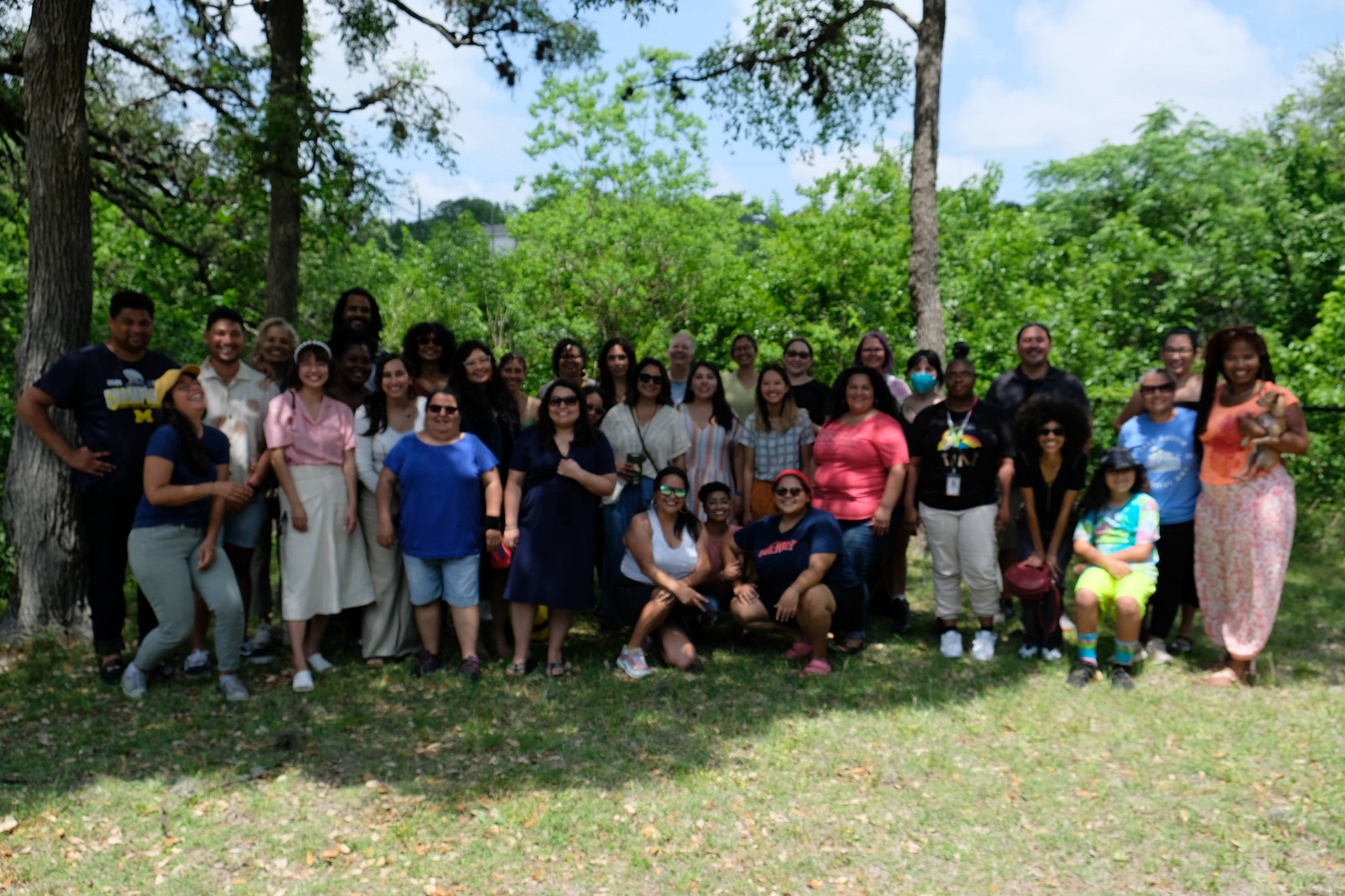 A group of diverse people from a previous EAT meeting stand outside in a park under trees and smile at the camera.