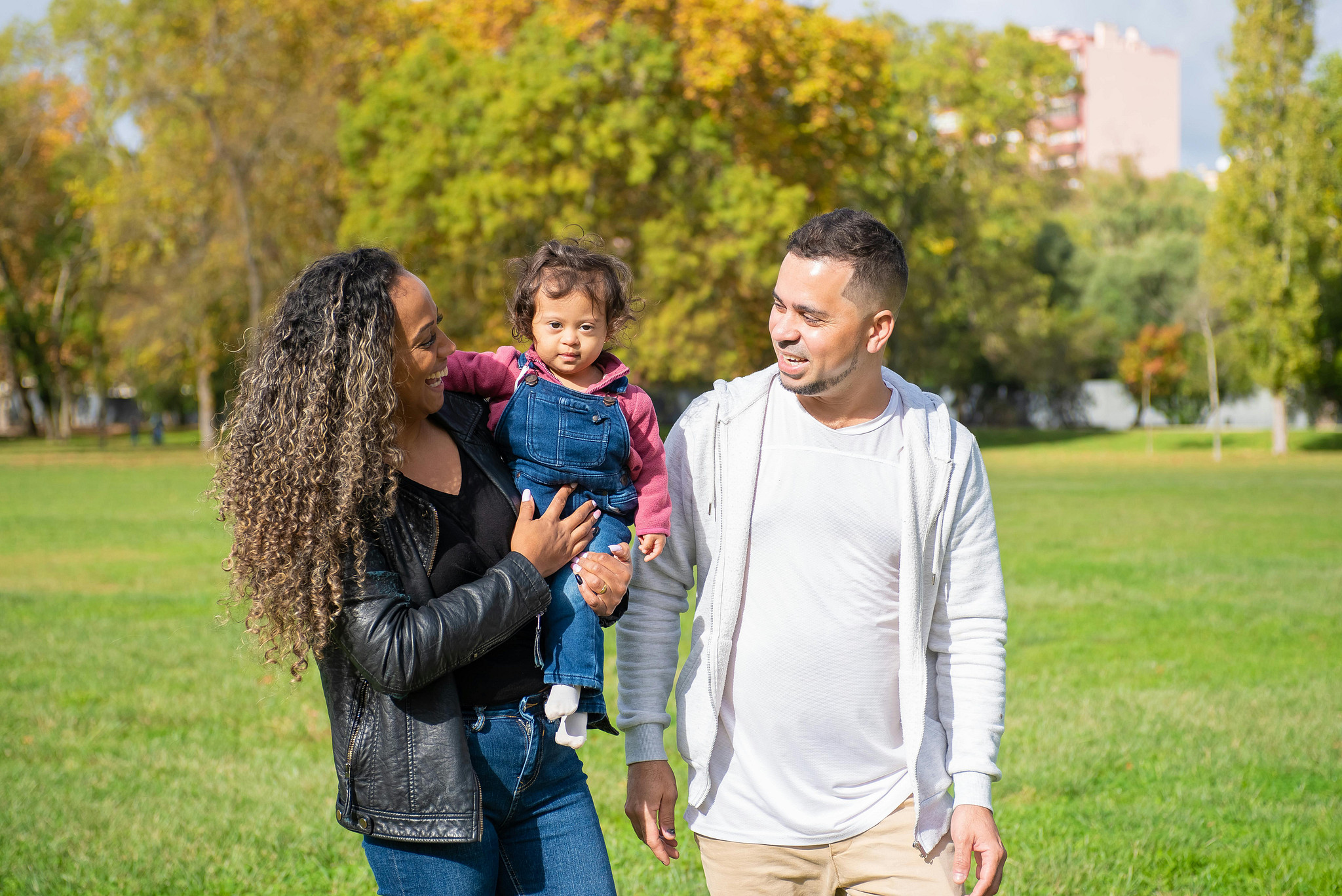 A multiracial family waks in a park with green field behind then. A woman of color with curly hair holds a toddler of color. A man of color smiles looking at the small girl.